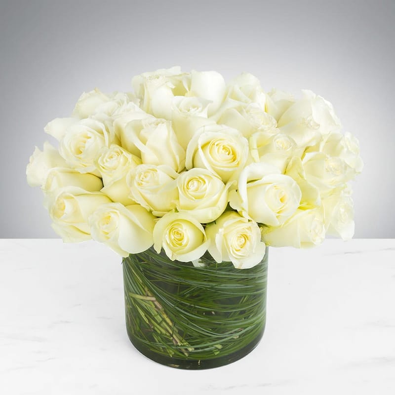 A dense bouquet of dozens of white roses in a clear glass cylinder vase. The inside of the vase is lined with blades of dark green grass, creating a modern look.