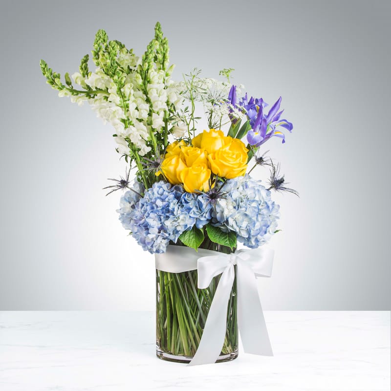 A bright and colorful Celebration of Life flower arrangement with blue hydrangeas, yellow roses, purple irises, and white snapdragons in a glass vase with a white bow.