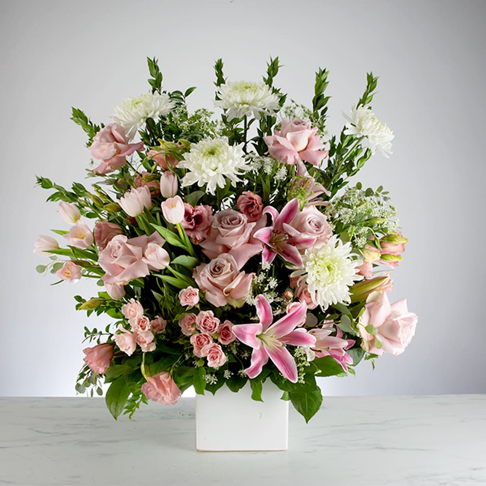 A large, elegant sympathy flower arrangement in a square white vase, featuring pink lilies, dusty mauve roses, white chrysanthemums, and pink tulips.