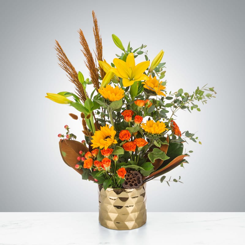 A tall autumn floral arrangement featuring yellow lilies, sunflowers, and orange spray roses with dried brown grasses, a lotus pod, and large magnolia leaves in a modern, geometric gold vase on a marble surface.