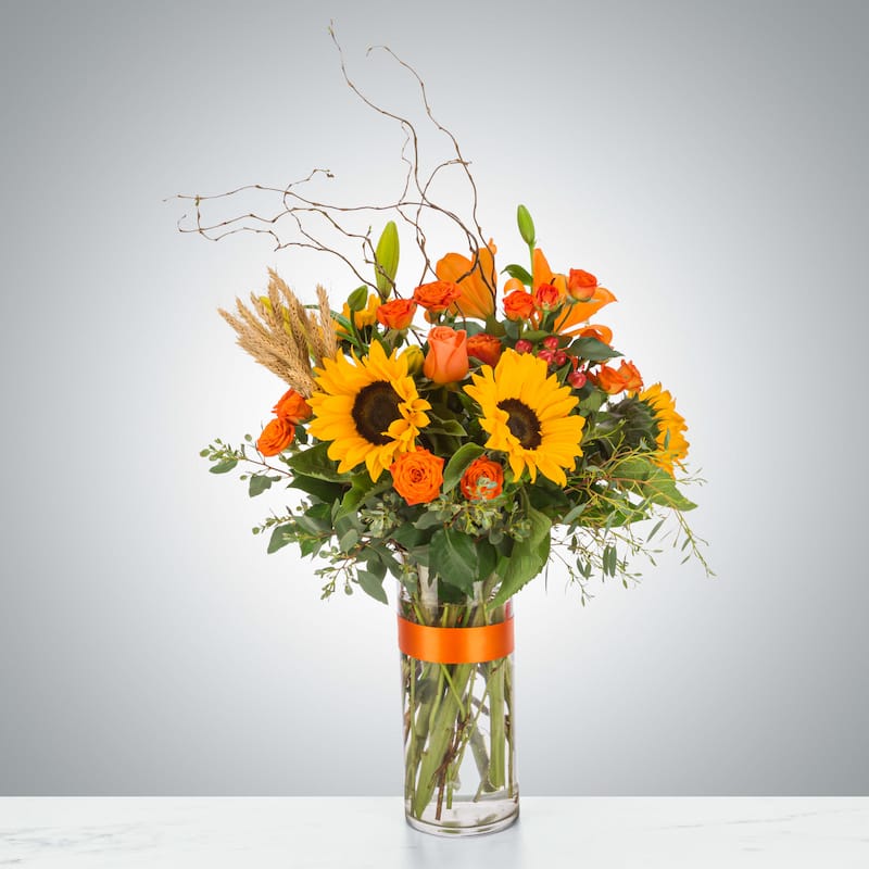 A classic autumn floral arrangement with sunflowers, orange lilies, and spray roses, accented with wheat stalks and curly willow branches in a clear glass vase tied with an orange ribbon.