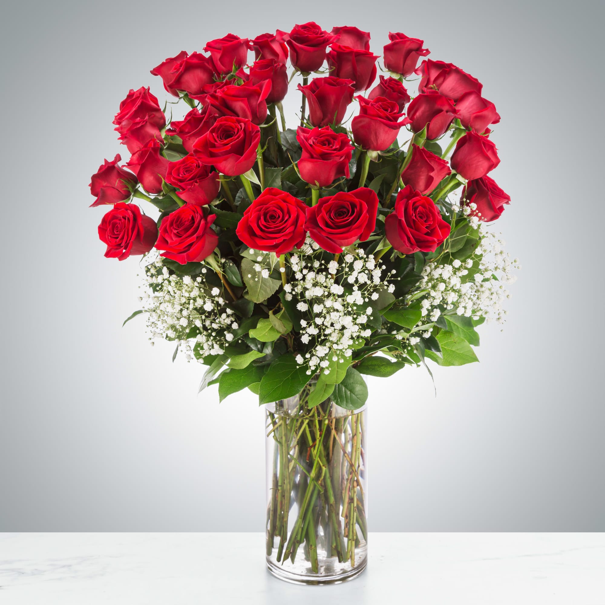 A luxurious floral arrangement named "Endless Devotion," featuring three dozen long-stemmed red roses and baby's breath elegantly displayed in a tall, clear glass vase on a white marble surface.