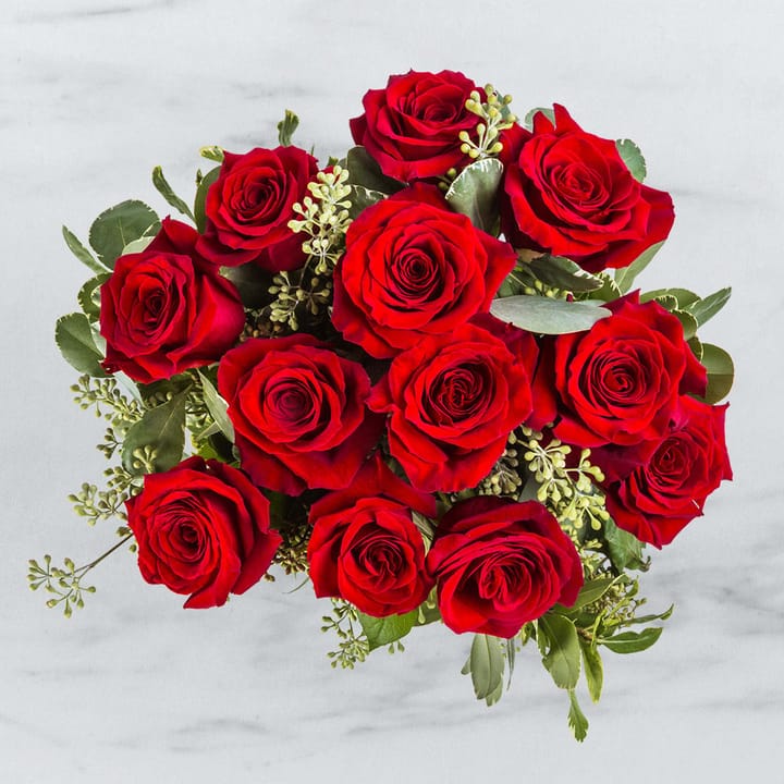An overhead view of a lush bouquet of a dozen vibrant red roses, artfully arranged with delicate green filler leaves, set against a white marble surface.