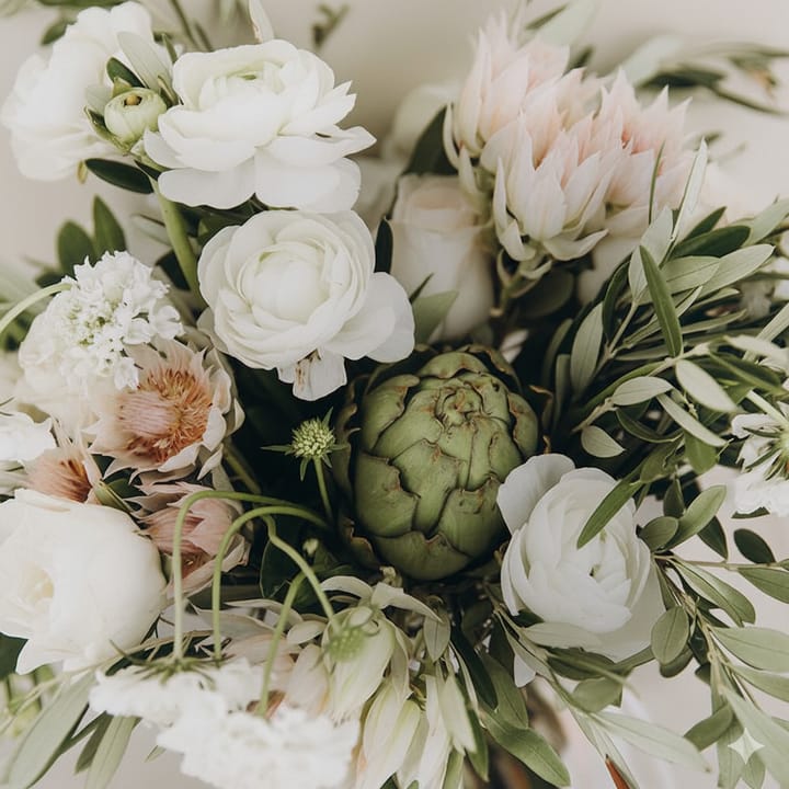 A close-up of a modern and textural sympathy arrangement featuring white ranunculus, blush pink protea, a green artichoke, and olive leaves.