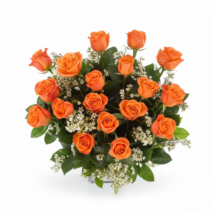 A professional floral arrangement of vibrant coral roses, delicate white wax flowers, and fresh green lemon leaf artfully designed in a clear glass vase, sitting on a clean white surface.