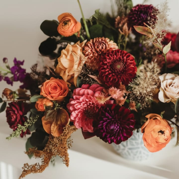 A lush autumn Thanksgiving centerpiece in a patterned ceramic vase, featuring rich burgundy dahlias, orange ranunculus, and textural dried grasses for a sophisticated holiday table.